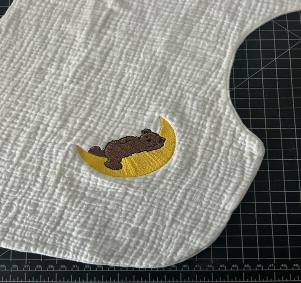 Baby bib with bear and moon embroidery on a textured white fabric.